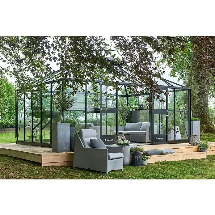 Juliana Gewächshaus Grand Oase - Außenmaß inkl. Dachüberstand (B x T): 511 x 368 cm, Material Seitenwand: Glas, 3 mm, Anthrazit - Schwarz gerahmtes Glasgewächshaus auf Holzdeck, als Gartenlounge mit Sitzgelegenheiten drinnen und draußen eingerichtet.