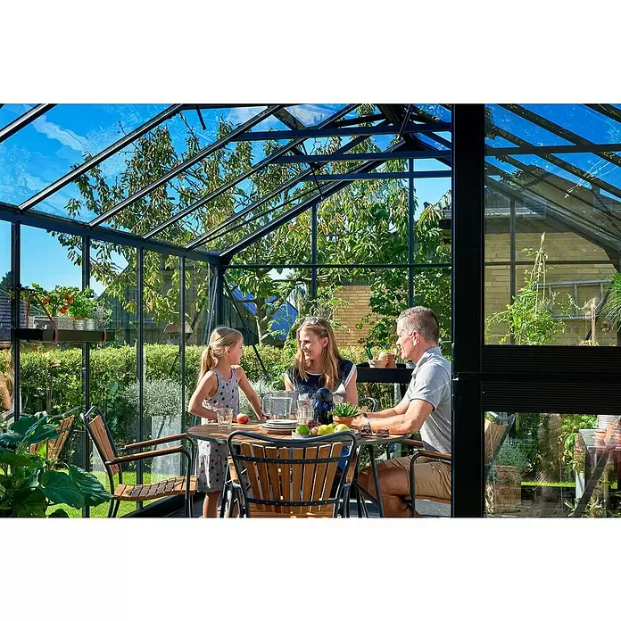 Juliana Gewächshaus Premium - Außenmaß inkl. Dachüberstand (B x T): 296 x 439 cm, 3 mm, Anthrazit, Einscheibensicherheitsglas - Modernes Glashaus mit schwarzem Rahmen, wo eine Familie an einem Tisch isst, umgeben von Gartenblick.