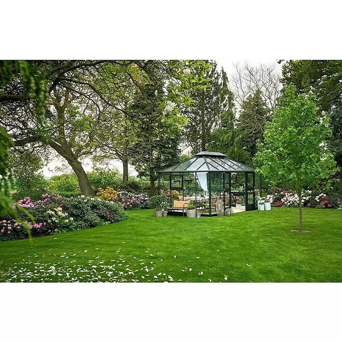 Juliana Gewächshaus Grand Oase - Außenmaß inkl. Dachüberstand (B x T): 439 x 296 cm - Gartenpavillon aus Glas mit schwarzem Rahmen und Esstisch, umgeben von grünem Rasen und blühenden Pflanzen.