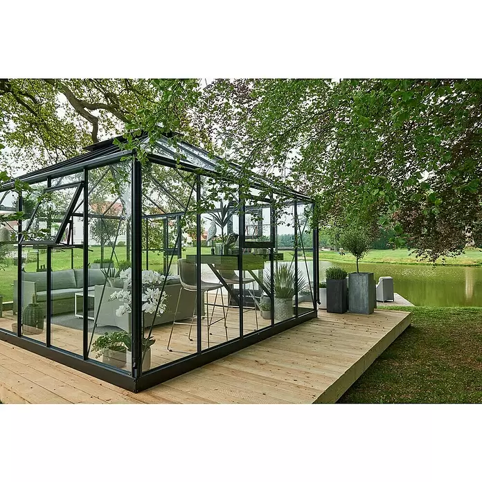 Juliana Gewächshaus Grand Oase - Außenmaß inkl. Dachüberstand (B x T): 511 x 368 cm, Material Seitenwand: Glas, 3 mm, Anthrazit - Schwarz gerahmtes Glasgewächshaus mit Sitzgelegenheiten und Pflanzen, auf einer Holzterrasse an einem ruhigen See.