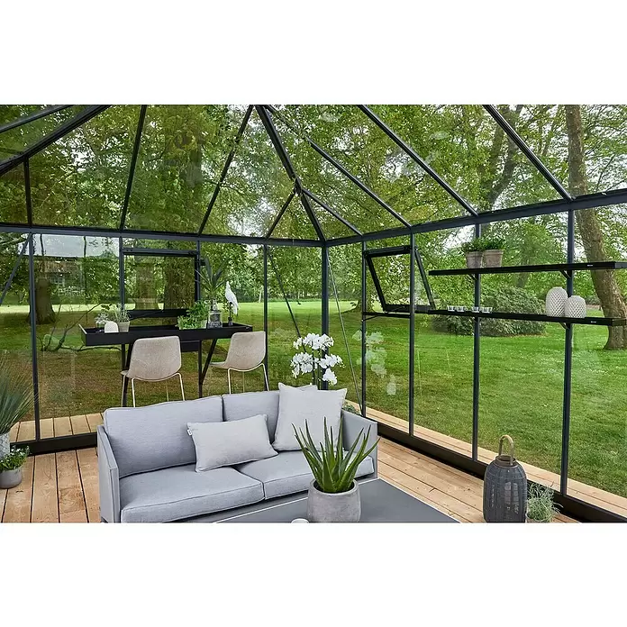 Juliana Gewächshaus Grand Oase - Außenmaß inkl. Dachüberstand (B x T): 511 x 368 cm, Material Seitenwand: Glas, 3 mm, Anthrazit - Geräumiges Glashaus mit dunklen Rahmen, eingerichtet als elegantes Wohnzimmer mit grauem Sofa und Essbereich, umgeben von Garten.