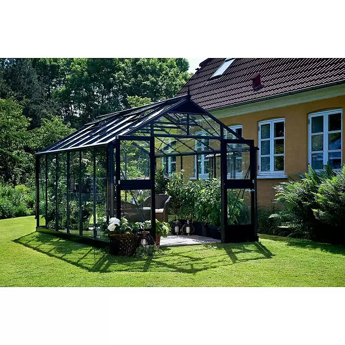 Juliana Gewächshaus Premium - Außenmaß inkl. Dachüberstand (B x T): 296 x 439 cm, 3 mm, Anthrazit, Einscheibensicherheitsglas - Großes Gewächshaus mit schwarzem Rahmen und Glasplatten in einem sonnigen Garten, für Pflanzen und Sitzgelegenheiten, neben einem gelben Haus.