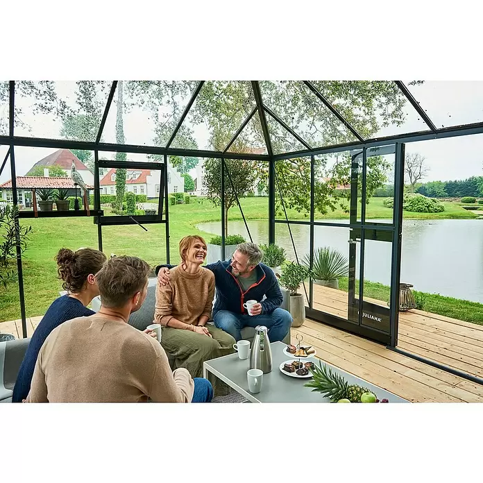 Juliana Gewächshaus Grand Oase - Außenmaß inkl. Dachüberstand (B x T): 511 x 368 cm, Material Seitenwand: Glas, 3 mm, Anthrazit - Vier Personen entspannen in einem modernen Juliana-Gewächshaus mit schwarzen Rahmen und Blick auf Garten und See.