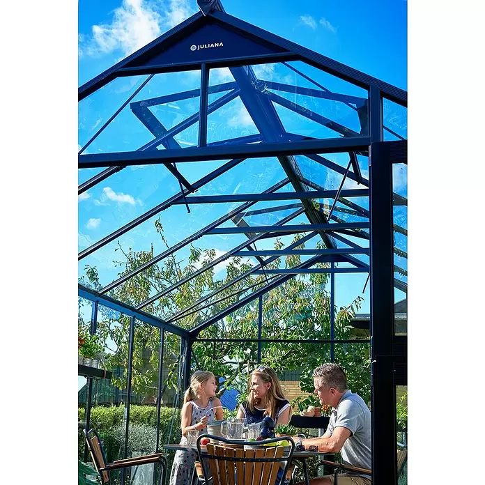 Juliana Gewächshaus Premium - Außenmaß inkl. Dachüberstand (B x T): 296 x 439 cm, 3 mm, Anthrazit, Einscheibensicherheitsglas - Juliana Gewächshaus aus dunkel gerahmtem Glas, in dem eine Familie eine Mahlzeit genießt.