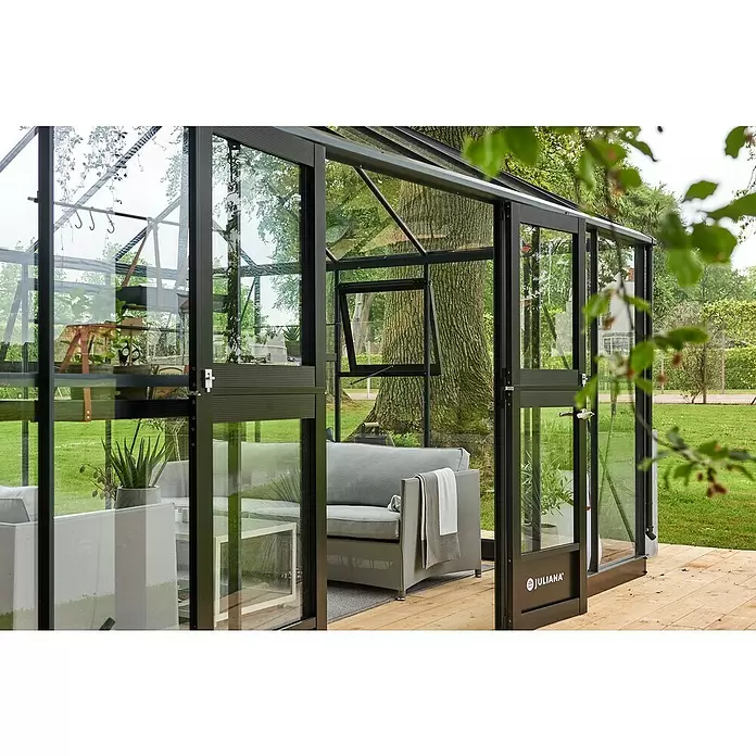 Juliana Gewächshaus Grand Oase - Außenmaß inkl. Dachüberstand (B x T): 511 x 368 cm, Material Seitenwand: Glas, 3 mm, Anthrazit - Dunkelgrau gerahmtes Glashaus als Gartenraum mit Sofa, auf Holzterrasse im Freien.