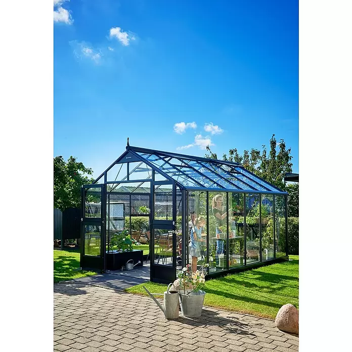Juliana Gewächshaus Premium - Außenmaß inkl. Dachüberstand (B x T): 296 x 439 cm, 3 mm, Anthrazit, Einscheibensicherheitsglas - Großes Gewächshaus mit schwarzem Rahmen und Glas im Garten, eine Familie pflegt Pflanzen darin.