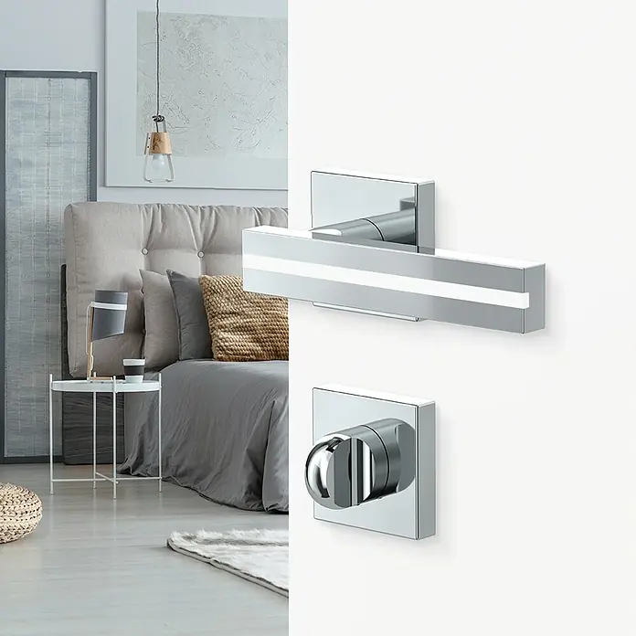 Diamond Doors Black Edition Zimmertürgarnitur WC Hampton White - Chrom, Stärke Tür: 40 mm - 45 mm, Schlitzkopf/Olive - Nahaufnahme eines modernen Türgriffs aus Chrom mit einem schlichten, rechteckigen Design vor einem stilvollen Schlafzimmerhintergrund.