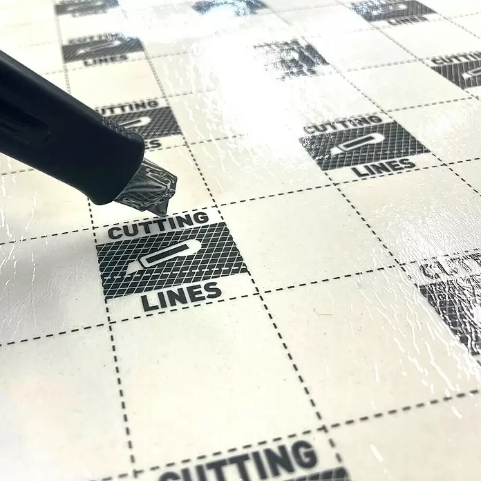 Base aislante - 9 m², Espesor: 1,3 mm - Un cúter negro con hoja metálica reposa sobre una alfombrilla de corte blanca con líneas de cuadrícula negras y las palabras "CUTTING LINES".