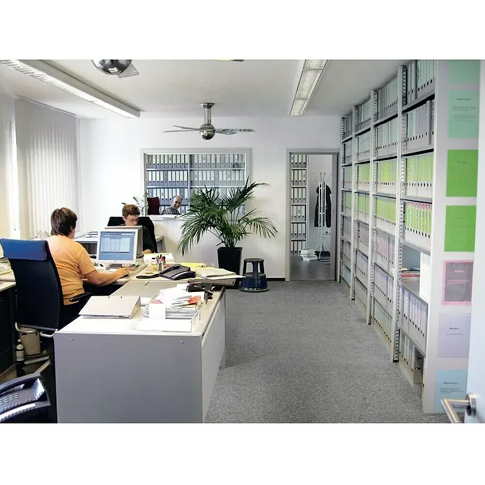 Scholz Aktenregal Grundelement - H x B x T: 264 x 105 x 30 cm, Traglast pro Boden: 80 kg, Anzahl Böden: 8 Stk., Stecken, Silber - Bürointerieur mit Büromöbeln und Regalen. Eine Person sitzt an einem Schreibtisch, während eine andere Person im Hintergrund arbeitet.