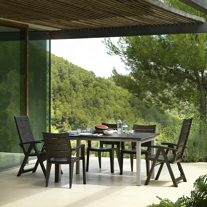 Silla de jardín Legno - Marrón, Polipropileno, Apilable, 60 x 59 x 87 cm - Escena exterior con un elegante juego de mesa de comedor. El conjunto consta de una mesa con patas plateadas y sillas a juego en marrón.