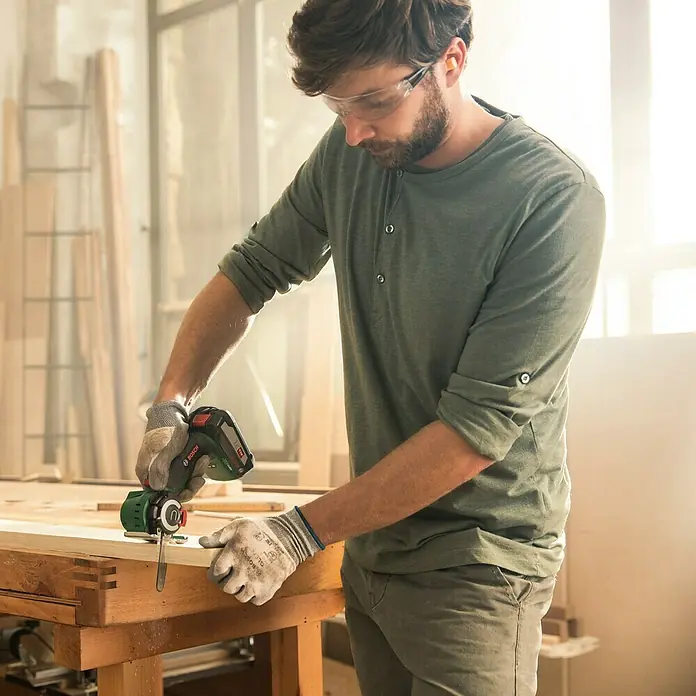 Bosch Power for All 18V NanoBlade-Säge AdvancedCut 18Leerlaufdrehzahl: 7.000 U/min, 18 V, Ohne Akku - Ein Mann benutzt ein Bosch Multifunktionswerkzeug, um an einem Holzstück zu arbeiten. Er trägt Schutzbrille und Handschuhe für die Arbeitssicherheit.