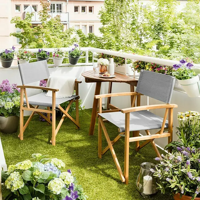 Sunfun Diana Balkontisch - L x B x H: 100 x 50 x 75 cm, Akazie, Klappbar, Gebogen, Naturbraun - Gemütlicher Balkon mit zwei hellgrauen Regiestühlen und einem kleinen Holztisch, umgeben von üppigen Blumenarrangements.