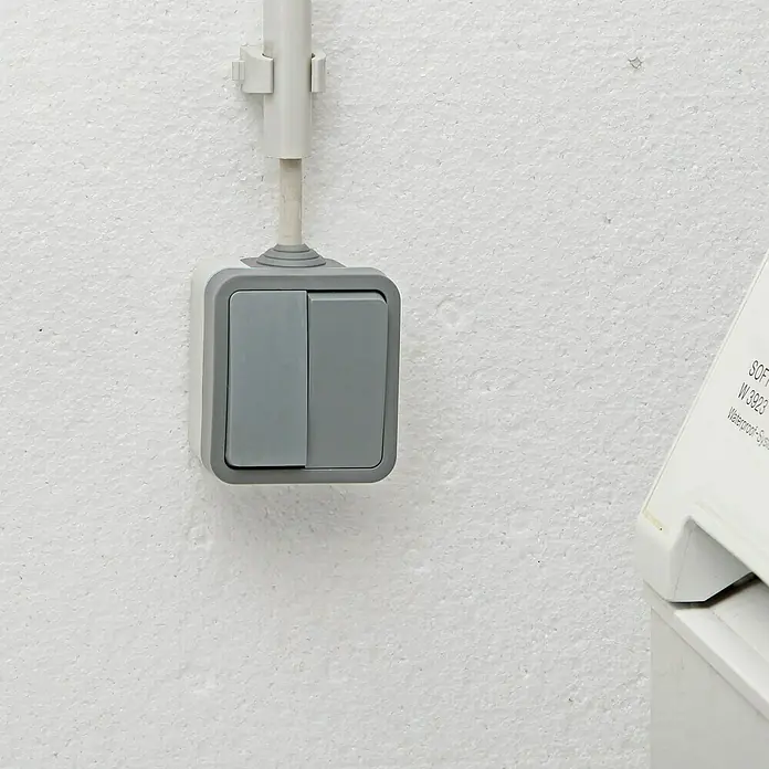 Voltomat Blueline Feuchtraum-Serienschalter - Grau, Aufputzmontage, IP54 - Nahaufnahme eines grauen Doppel-Lichtschalters an einer weißen Wand mit Kabel und Halterung.