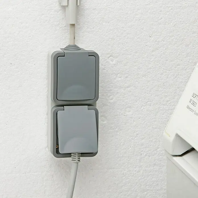 Voltomat Blueline Feuchtraum-Steckdose - 2-fach, Vertikal, IP54, Grau - Nahaufnahme einer grauen Doppelsteckdose mit hochgeklappten Deckeln, die an einer weißen Wand montiert ist.