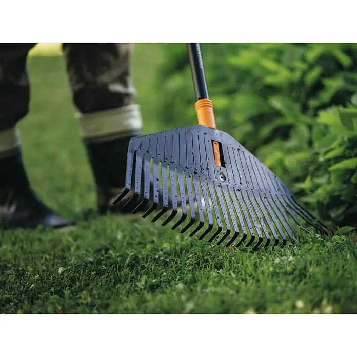 Fiskars Solid Laubbesen - Arbeitsbreite: 52 cm, Anzahl Zinken: 24, Art Stiel: Langstiel - Nahaufnahme eines schwarzen Laubrechens auf Rasen, mit einer Person im Hintergrund. Ideal für Gartenarbeit und Landschaftspflege.