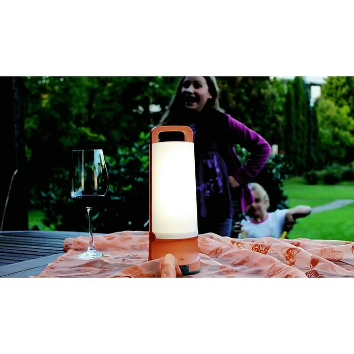 Lutec Lámpara decorativa solar LED Dragonfly - L x An x Al: 10,2 x 10,2 x 28,2 cm, Naranja, IP54 - Primer plano de una lámpara portátil junto a una copa de vino sobre un mantel, con niños jugando al fondo.