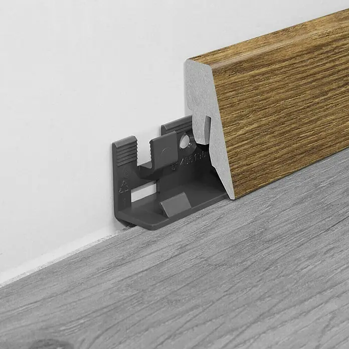 LOGOCLIC Sockelleiste K40 C Eiche Mirandola - 260 cm x 19 mm x 40 mm, MDF - Sockelleisten-Clip zur Befestigung einer braunen Holzsockelleiste an einer weißen Wand über grauem Boden.