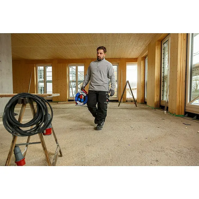 Brennenstuhl Kabeltrommel Garant CEE 1 - Kunststoff, 30 m, H07RN-F, IP44 - Ein Handwerker in einem Rohbau, der einen blauen Kabeltrommel trägt, neben einer  auf einem Bock liegenden, schwarzen Stromkabelrolle.