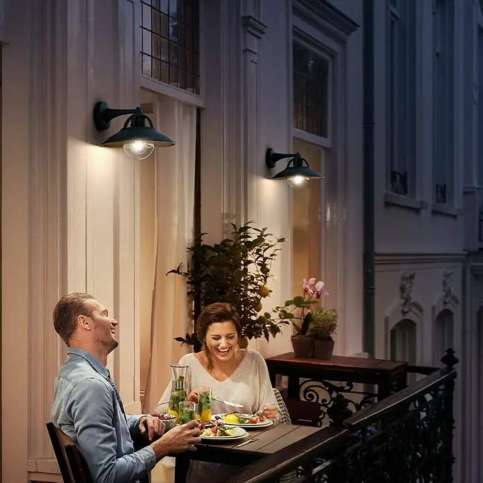 Philips Aplique exterior Cormorant - 42 W, 265 x 250 x 218 mm, Negro, IP44 - Una elegante luz exterior ilumina un patio donde una pareja disfruta de la cena. Las luces crean un ambiente exterior acogedor.