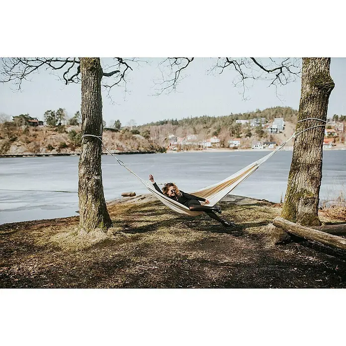 Amazonas Hängematte Barbados - L x B: 230 x 150 cm, Textil, Beige - Eine Frau entspannt in einer Hängematte, die zwischen zwei Bäumen gespannt ist, mit Blick auf einen ruhigen See in einer friedlichen Umgebung.
