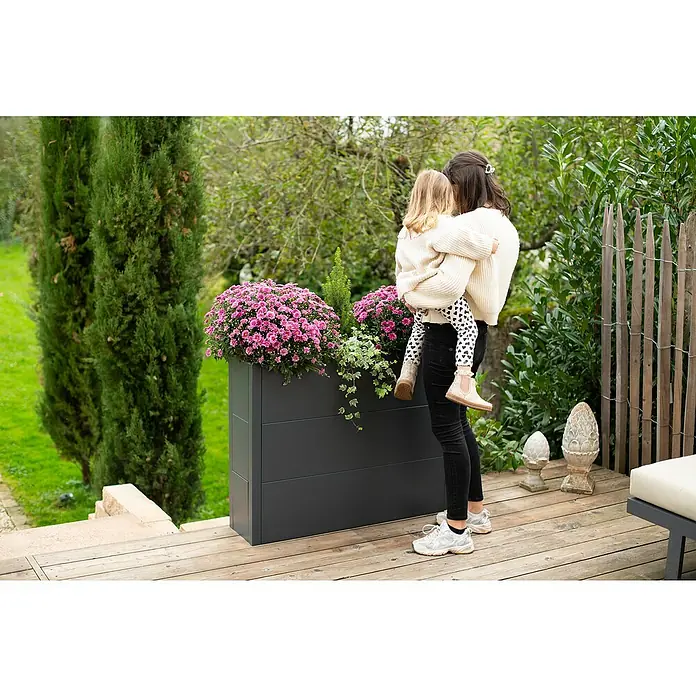 Westmann Pflanzkasten Talus XL - L x B x H: 25 x 100 x 77 cm, Metall, Anthrazit - Eine Mutter hält ihr Kind vor einem grauen Pflanzkasten mit rosa Blumen auf einer Holzterrasse.