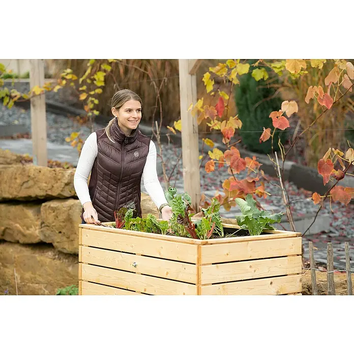 Westmann Hochbeet Asket M - 70 x 110 x 90 cm, Holz, Natur - Frau im Garten mit einem Hochbeet aus Holz, in dem verschiedene Pflanzen wachsen, während sie lächelt.