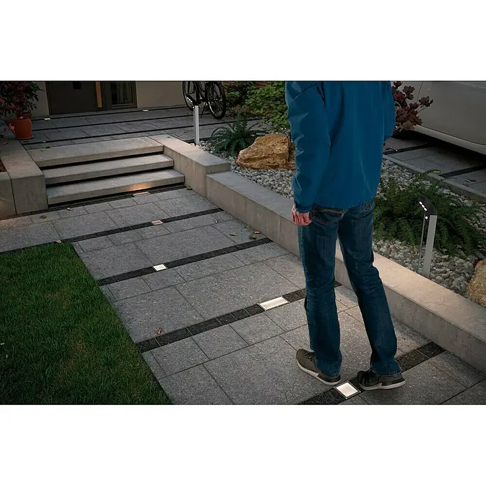 Paulmann Foco para empotrar en suelo Aron - 1,5 W, Blanco, L x An: 10 x 20 cm - Imagen de iluminación exterior empotrada en el suelo que ilumina un camino mientras una persona camina sobre él.