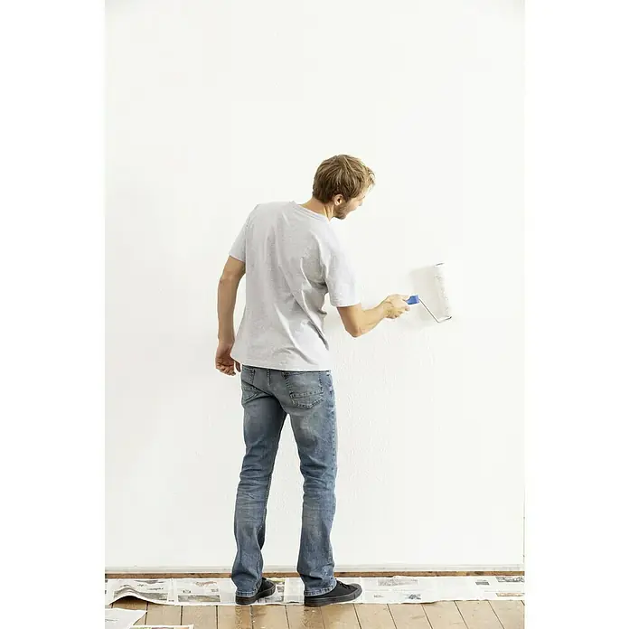 Knauf EasyPutz Finishwalze - Geeignet für: Putz, Breite Walze: 22 cm, Bügelstärke: 8 mm - Ein Mann streicht eine Wand mit einer weißen Farbwalze. Er trägt Jeans und ein graues T-Shirt und die Zeitung schützt den Boden.