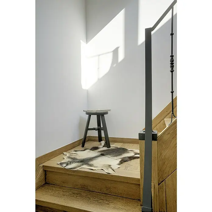 Esbeco Ziegenfell - Natur, 100 % Echtfell, 70 x 55 cm - Einrichtungselement: Hocker mit Fellauflage im Treppenhaus, beleuchtet durch einfallendes Sonnenlicht. Stilvolles Detail für den Wohnbereich.