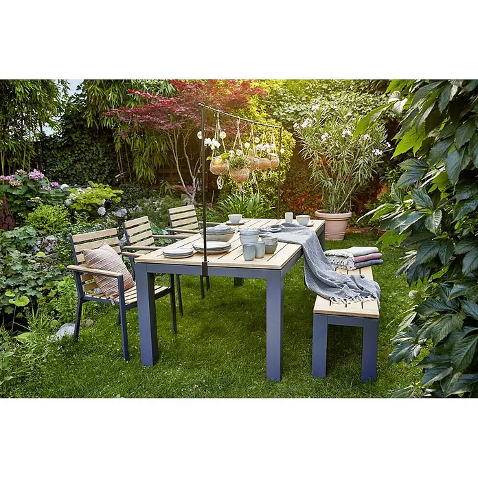Sunfun Carina Gartenstuhl - Akazie, Stapelbar, Natur, B x T x H: 57,5 x 61 x 86 cm - Ein Gartentisch-Set mit Stühlen und Bank auf einer grünen Rasenfläche, umgeben von üppigem Grün. Dekorative Hängetöpfe hängen über dem Tisch.