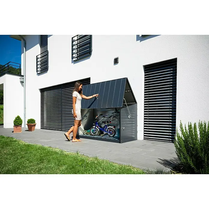 Trimetals Garten-Aufbewahrungsbox Storeguard - Anthrazit, Stahl - Eine Frau öffnet einen Fahrradschuppen aus Metall, in dem sich ein Fahrrad und ein Roller befinden. Der Schuppen steht an einer modernen Hauswand.