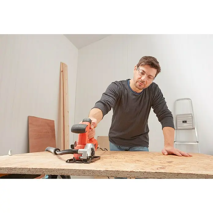 Black+Decker Minisierra circular de mano BES510-QS - 400 W, Diámetro hoja de sierra: 85 mm, Número de revoluciones de marcha en vacío: 3.600 r.p.m. - Un hombre usa una sierra circular BLACK+DECKER para cortar una tabla de madera. Lleva gafas de seguridad. Una escalera está en el fondo.