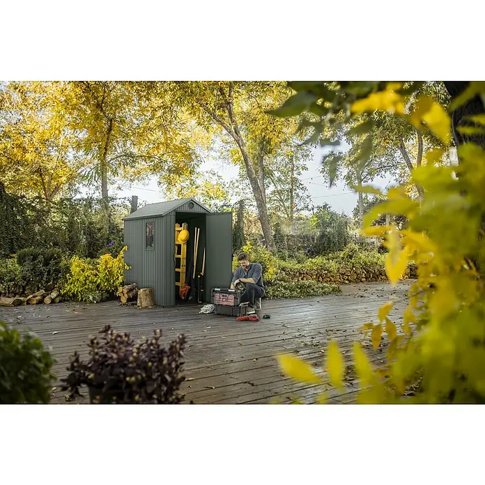 Keter Gerätehaus Darwin 4x6 - Außenmaß inkl. Dachüberstand (B x T): 125,8 x 184,5 cm, Kunststoff, Grün - Dunkelgraues Gartenhaus mit geöffneter Tür und Mann davor, ideal für die Aufbewahrung von Gartengeräten und Werkzeugen.
