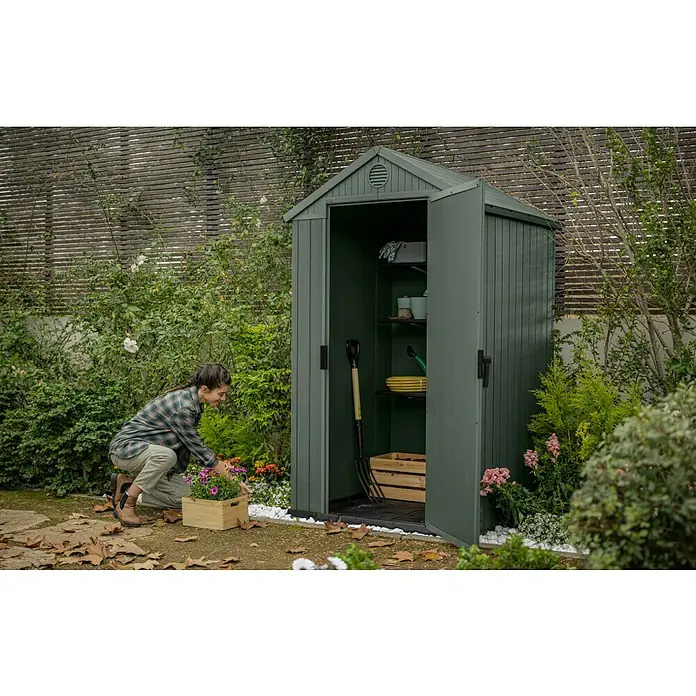 Keter Gartenhaus Darwin 4x4 - Außenmaß inkl. Dachüberstand (B x T): 125,8 x 117 cm, Kunststoff, Grün, 1,5 m² - Ein geräumiger, grüner Geräteschuppen im Garten mit geöffneten Türen, der Gartenwerkzeuge und Regale zeigt, während eine Frau im Garten arbeitet.