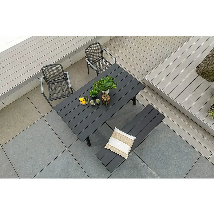 Sunfun Melina Gartenbank - B x T x H: 155 x 42,4 x 45,7 cm, Akazie, Black Washed - Ein Gartentisch-Set mit zwei Stühlen und einer Bank auf einer gepflasterten Terrasse. Der Tisch ist mit Pflanzen und Geschirr dekoriert.