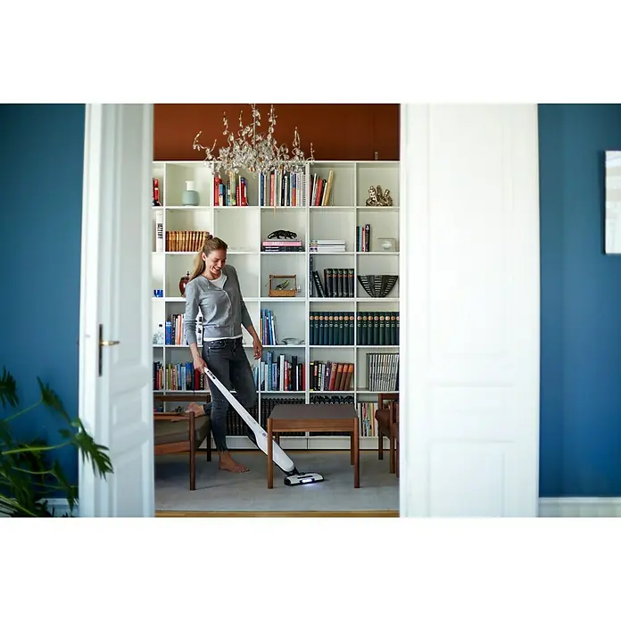 Nilfisk Akku-Staubsauger Easy -  - Frau benutzt einen weißen Philips Staubsauger in einem stilvollen Raum mit Bücherregal. Praktische Reinigung leicht gemacht.