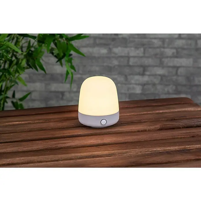 Starlux Akku-Tischleuchte Mini - L x B x H: 10 x 10 x 12 cm, Beige, RGBW, 0,3 W, IP44 - Eine Nachttischlampe mit sanftem, warmem Licht steht auf einem Holztisch und sorgt für eine gemütliche Atmosphäre.
