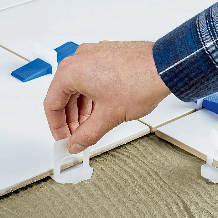 Heka Nivofix Nivelliersystem201 -tlg. - Bild einer Hand, die ein Nivelliersystem für Fliesen anbringt, mit weißen Fliesen und blauem Keilen für präzise Installationen.