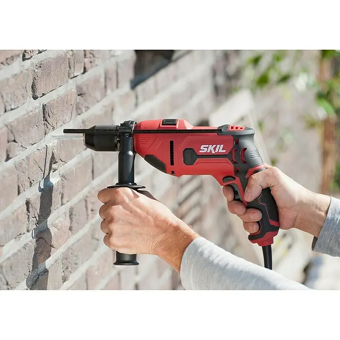 Skil Schlagbohrmaschine 6725 AA710 W, Schnellspannbohrfutter - Eine rote SKIL Bohrmaschine bohrt ein Loch in eine Ziegelwand, gehalten von zwei Händen, für Heimwerker und Bauprojekte.