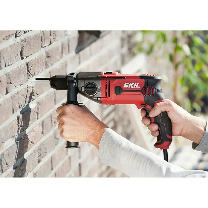 Skil Schlagbohrmaschine 6745 GA - 1.050 W, Schnellspannbohrfutter - Ein roter SKIL Hammerbohrer wird zum Bohren in eine Ziegelwand verwendet, was die Leistungsfähigkeit und Vielseitigkeit des Werkzeugs hervorhebt.