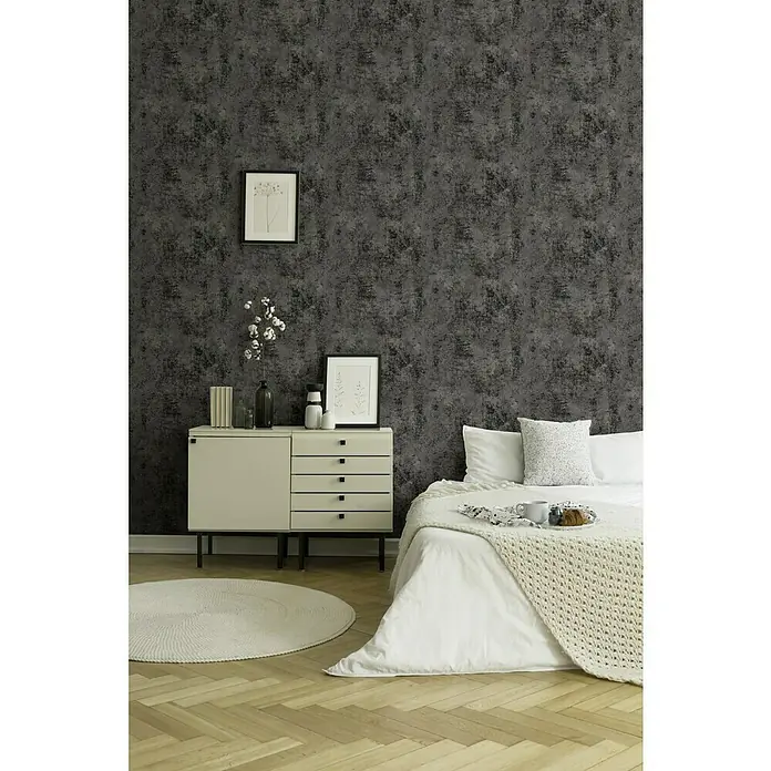 A.S. Création New Walls Vliestapete - Anthrazit, Uni, 10,05 x 0,53 m - Aufnahme eines Schlafzimmers mit grauer gemusterter Tapete, hellem Nachttisch und Bett mit weißer Bettwäsche.
