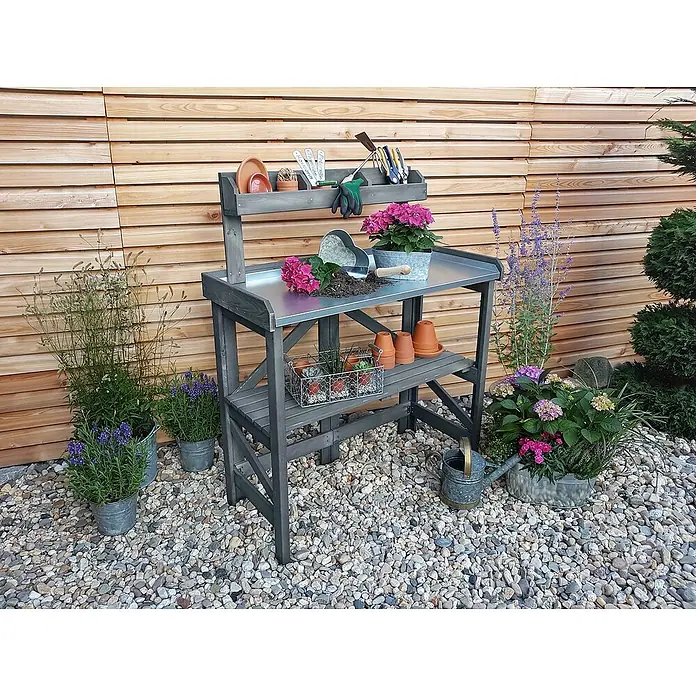 Promadino Pflanztisch Klappbar - 97 x 45 x 128 cm, Holz, Grau - Ein grauer Gartentisch mit Blumen, Töpfen und Gartengeräten vor einer Holzwand auf Kies zeigt die Funktionalität und Ästhetik.