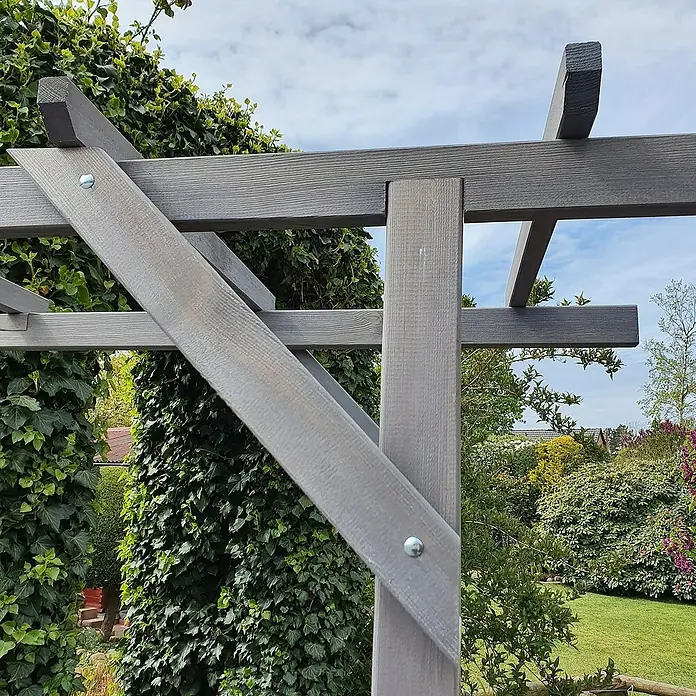 Promadino Pergola Ben - 60 x 200 x 210 cm, Grau, Holz - Nahaufnahme einer grau gestrichenen Holzpergola mit Schrauben, umgeben von üppigem Grün und einem blauen Himmel.