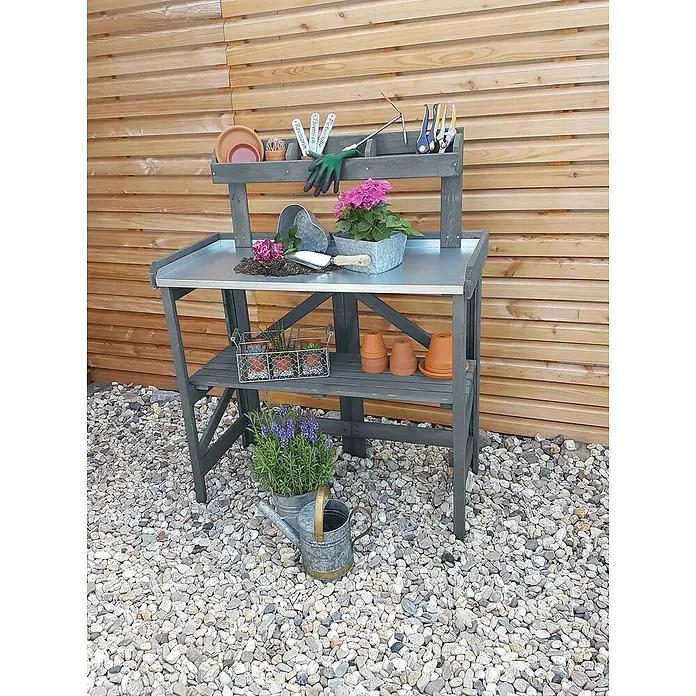Promadino Pflanztisch Klappbar - 97 x 45 x 128 cm, Holz, Grau - Eine graue Gartenwerkbank mit Pflanzen und Werkzeugen darauf, die auf einem Kiesweg vor einer Holzwand steht.