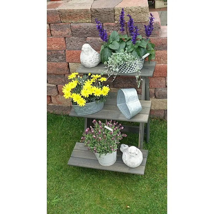 Promadino Blumentreppe - 60 x 50 x 56 cm, Kiefer, Grau - Dreistufiges Regal mit Blumen und dekorativen Vogelstatuen auf einer grünen Rasenfläche vor einer Ziegelmauer.
