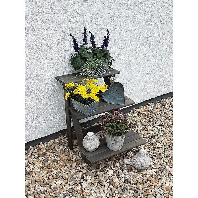 Promadino Blumentreppe - 60 x 50 x 56 cm, Kiefer, Grau - Dreistufiges Holzregal mit Blumen in Töpfen und dekorativen Vogelstatuen, platziert auf einem Kiesbett vor einer weißen Wand.