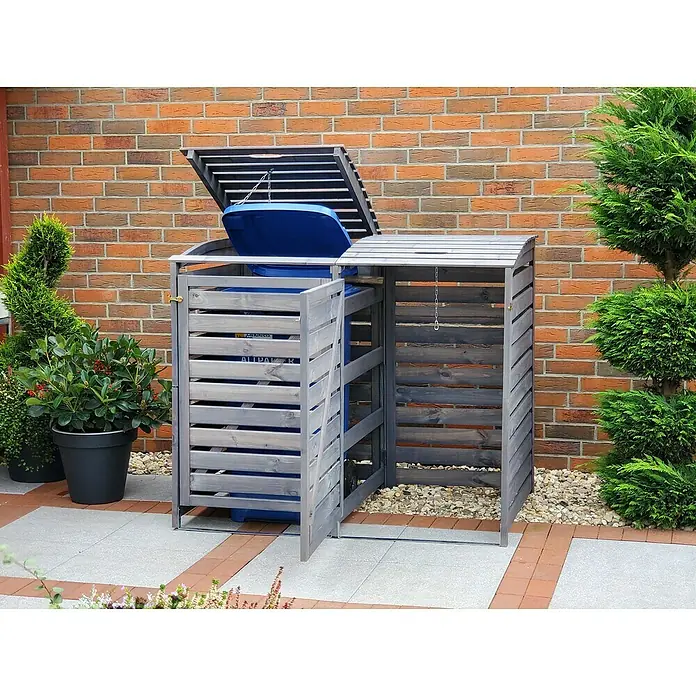 Promadino Mülltonnenbox Vario III -  - Eine Doppel-Mülltonnenbox aus Holz steht im Garten vor einer Backsteinmauer, mit Pflanzen drumherum.