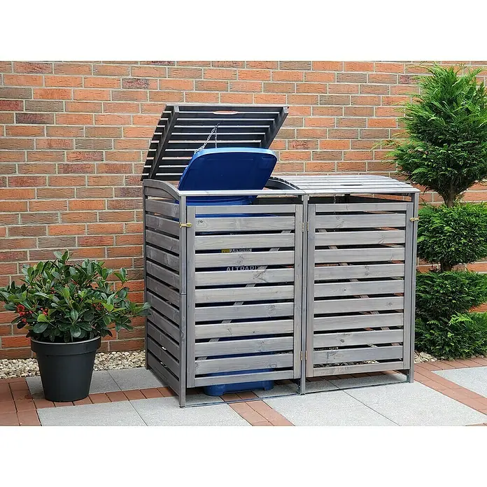 Promadino Mülltonnenbox Vario III -  - Grauer, hölzerner Mülltonnenbox mit zwei Fächern, Deckel teilweise geöffnet, vor einer Ziegelmauer und Grünpflanzen aufgestellt.