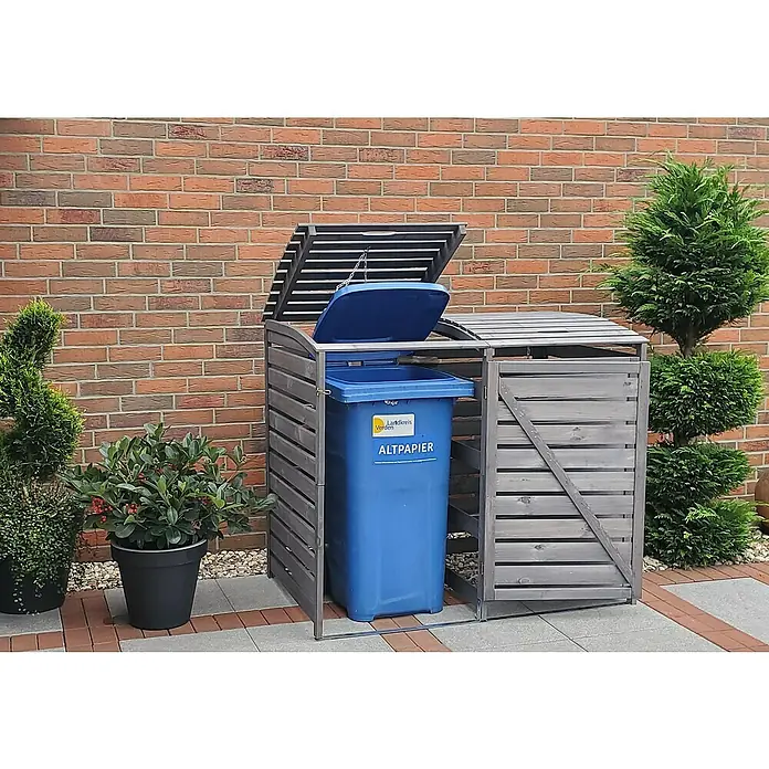 Promadino Mülltonnenbox Vario III -  - Eine graue Mülltonnenbox aus Holz mit geöffnetem Deckel und einer blauen Papiermülltonne davor, umgeben von Pflanzen und einer Ziegelmauer.