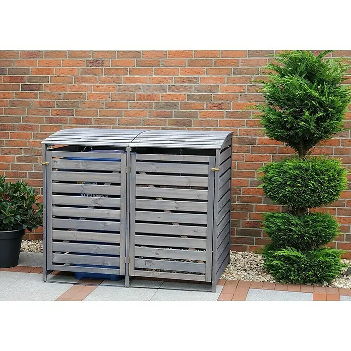 Promadino Mülltonnenbox Vario III -  - Das Bild zeigt einen doppelten Mülltonnenbox aus grauem Holz vor einer Ziegelmauer, neben einem Zierbaum und einem Topf mit Pflanzen.